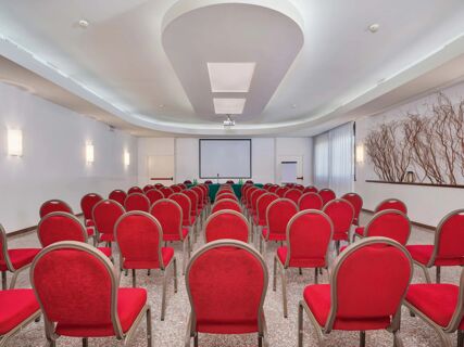 Conference room with multiple rows of red upholstered chairs facing a white screen and presentation table at B&B Hotel Eurorest Conegliano