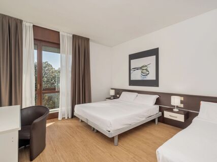 Guest room at B&B Hotel Eurorest Conegliano with two beds, wooden floor, nightstands, lamps, framed artwork, and a window with curtains