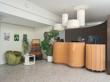 Reception area at B&B Hotel Eurorest Conegliano with wooden curved desk, hanging lamps, green chairs, plants, and stacked vintage suitcases