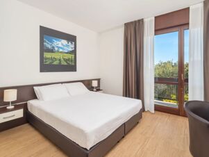 Bed with white linens, two bedside tables with lamps, vineyard artwork, and glass doors with brown curtains at B&B Hotel Eurorest Conegliano