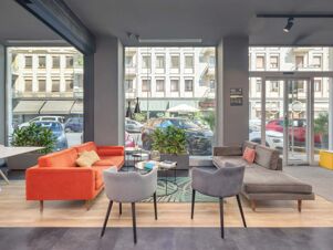 Interior lounge of B&B Hotel Milano Central Station with orange and gray sofas, armchairs, plants, and large street-facing windows