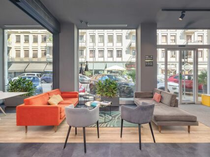 Interior lounge of B&B Hotel Milano Central Station with orange and gray sofas, armchairs, plants, and large street-facing windows