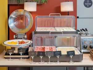 Buffet breakfast setup with scrambled eggs, bacon, cold cuts, sliced cheese, and sandwiches at B&B Hotel Milano Central Station