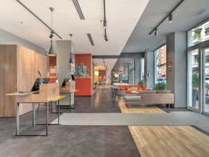 Modern hotel reception and seating area with wooden desks, pendant lights, gray chairs, and large windows at B&B Hotel Milano Central Station