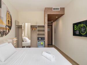 Modern hotel room at B&B Hotel Milano Central Station with white bedding, open wardrobe, wall TV, and wall art of city buildings