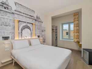 Bed with white linens beneath a large wall mural of an archway with yellow columns, window with polka dot yellow curtains in B&B Hotel Milano Central Station room
