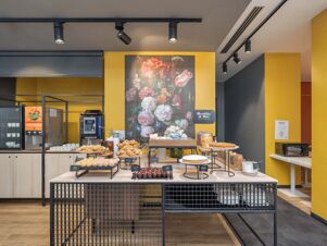 Breakfast buffet table with croissants, muffins, cereals, and coffee machine against a yellow wall at B&B Hotel Milano Central Station