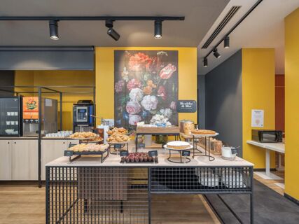 Breakfast buffet table with croissants, muffins, cereals, and coffee machine against a yellow wall at B&B Hotel Milano Central Station