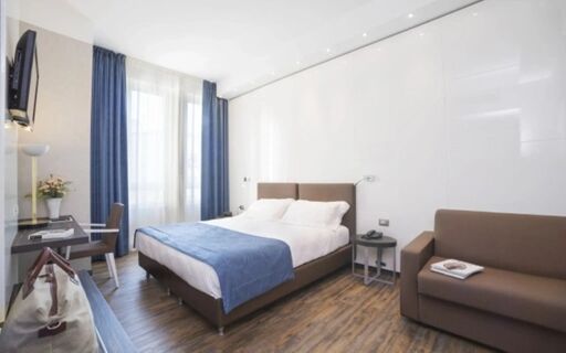 Hotel Atlantic room with a double bed covered by a blue blanket, brown sofa, desk with chair, and curtains on the window