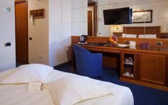 Hotel Berna room with white bed linens, blue chair, wooden desk, wall-mounted TV, and a fruit basket on the desk