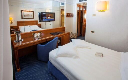 Hotel Berna room featuring a bed with white linens, blue carpet, wooden desk, blue chair, wall-mounted TV, and mirror panels