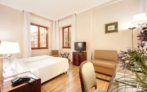 Hotel Bristol room with two beds, wooden flooring, a small sofa, TV on a dresser, desk with chair, and two wooden-framed windows