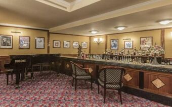 A classic bar area with patterned carpet, wooden chairs, a Bosoni grand piano, and framed artwork at Doria Grand Hotel