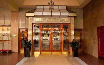 Entrance of Doria Grand Hotel with glass double doors, stone walls, two large potted plants, and a luggage cart on the left side