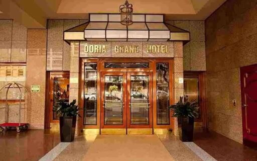 Entrance of Doria Grand Hotel with glass double doors, stone walls, two large potted plants, and a luggage cart on the left side