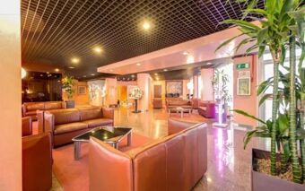Spacious lobby area of Hotel Galileo with brown leather sofas, glass-top tables, potted plants, and warm lighting
