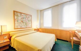 Hotel Galileo room with a double bed covered in beige bedding, green carpet, wooden furniture, and large windows with blinds