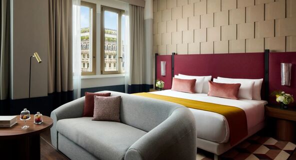 Hotel room at Tivoli Tivoli President Milano Hotel with a double bed, yellow throw, two pink pillows, gray sofa, and window view of a building