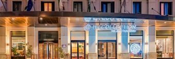 Exterior view of the illuminated entrance and glass windows of Hotel NH Collection Milano Touring with flags above and modern decor visible inside