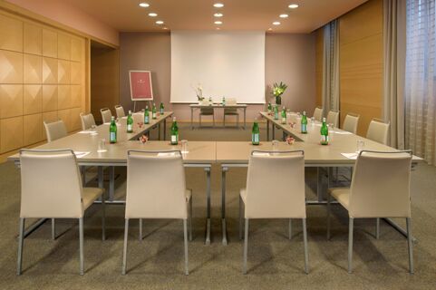 Conference room with U-shaped table setup, beige chairs, bottled water, bowls of snacks, and a projection screen at Hotel NH Collection Milano Touring