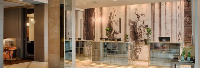 Reception area of Hotel NH Collection Milano Touring with modern mirrored desks, chairs, plants, and a wall featuring classical statues