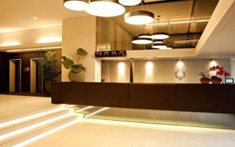 Hotel Manin reception area with modern brown counter, illuminated stairs, hanging circular lights, and digital time display showing world cities