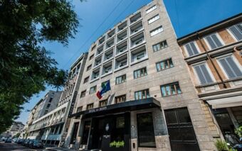 Hotel Manin entrance with flags mounted above, surrounded by a stone modern building facade and parked cars on the street