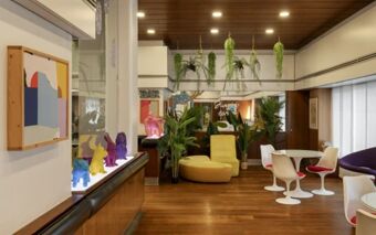 Lounge area at Hotel Mediolanum featuring wooden floors, modern chairs, a yellow sofa, plants, and colorful figurines on display