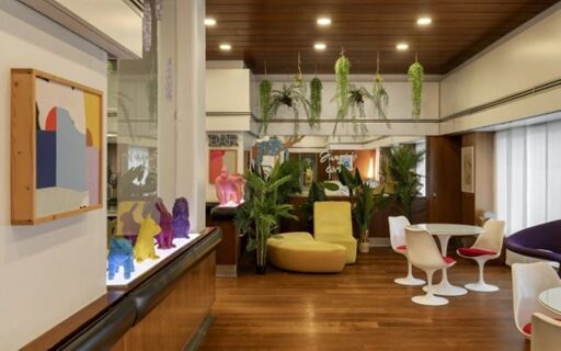 Lounge area at Hotel Mediolanum featuring wooden floors, modern chairs, a yellow sofa, plants, and colorful figurines on display