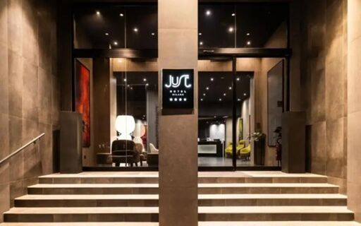 Entrance of Just Hotel Milano featuring wide stairs leading to glass doors framed by stone walls and illuminated signage