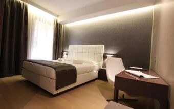 Hotel Baviera room with a white upholstered bed, brown throw, dark curtains, bedside lamps, and a desk with a chair and magazine