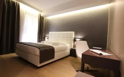 Hotel Baviera room with a white upholstered bed, brown throw, dark curtains, bedside lamps, and a desk with a chair and magazine