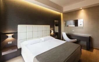 Hotel Baviera room featuring a neatly made bed with a white padded headboard, bedside lamps, a desk with a chair, and a wall mirror