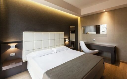 Hotel Baviera room featuring a neatly made bed with a white padded headboard, bedside lamps, a desk with a chair, and a wall mirror