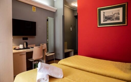 Twin beds with yellow bedspreads and a rolled towel, a red accent wall, TV, desk, and mirror in Hotel Berlino room