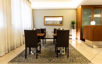 Dining area at Hotel Berlino with wooden tables, dark chairs, a patterned rug, white curtains, and a potted plant