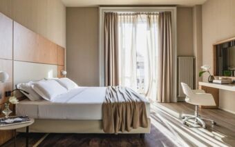 A bright hotel room at Hotel Cristoforo Colombo with a large bed, beige curtains, a desk, chair, and a wall-mounted TV