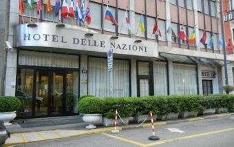 Facade of Hotel Delle Nazioni featuring multiple national flags above glass doors and rows of green shrubs along the entrance