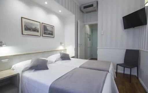 Two single beds with white linens and gray runners in a minimalistic room at Hotel Flora with a wall-mounted TV and chair