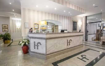 Marble reception desk with Hotel Flora logo in front of a well-lit lobby featuring plants and framed artwork on walls