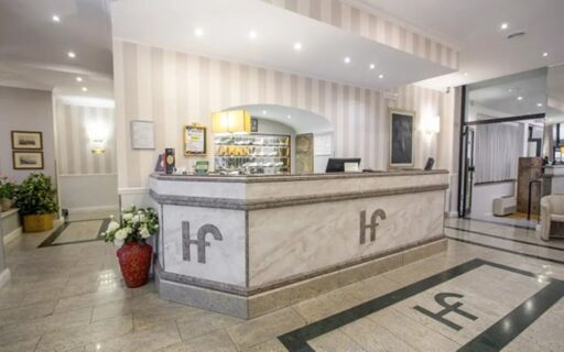 Marble reception desk with Hotel Flora logo in front of a well-lit lobby featuring plants and framed artwork on walls