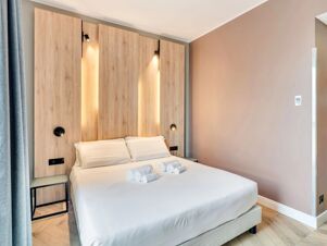 A neatly made double bed with four rolled white towels and wood-paneled headboard with built-in lights inside B&B Hotel Milano Aosta