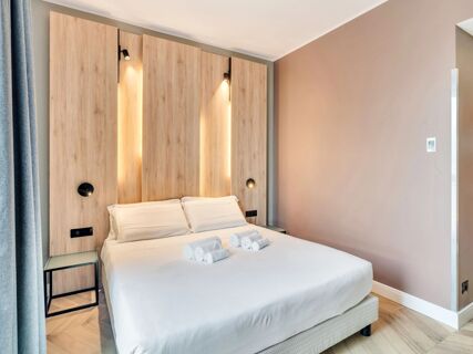 A neatly made double bed with four rolled white towels and wood-paneled headboard with built-in lights inside B&B Hotel Milano Aosta