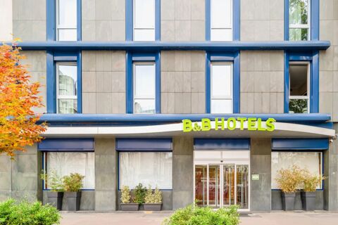 Exterior facade of B&B Hotel Milano Aosta featuring large windows framed in blue and an entrance with glass doors and plants nearby