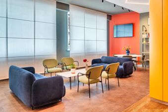 Modern lounge area with blue sofas, green chairs, a white table with plants, and a wall-mounted TV at B&B Hotel Milano Aosta