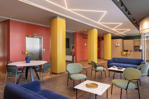 Lobby of B&B Hotel Milano Aosta with yellow pillars, red walls, modern ceiling lights, elevators, and seating areas with blue and green chairs