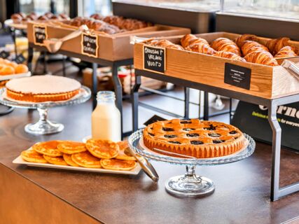 A breakfast buffet with croissants, pancakes, cakes, and milk displayed on a counter at B&B Hotel Milano Aosta