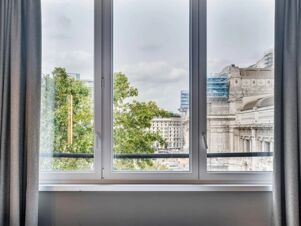 View through closed window with grey curtains showing green trees and historic buildings outside at B&B Hotel Milano Aosta