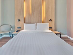 A double bed with white linens and pillows against a light wood headboard with vertical inset lighting and bedside tables at B&B Hotel Milano Portello