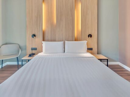 A double bed with white linens and pillows against a light wood headboard with vertical inset lighting and bedside tables at B&B Hotel Milano Portello